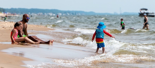 A smiling man and little girl sit on the wet, sandy beach, watching a toddler in sunhat and life vest walk in the low, blue-green waves 