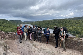 a small group of men and women in hiking gear and full backpacks stand next to a tan, rocky wall overlooking a blue river and lush green forest