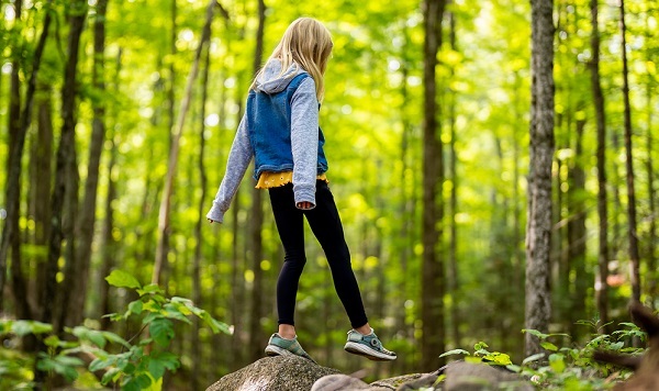 girl in black tights and gray-blue jacket and sneakers walks from one rock to another in a sunlit, lush green forest