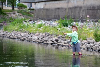 Blond woman in pale green shirt, sunglasses and tan hat stands knee-deep in a stream, holding a fly-fishing rod that's been cast