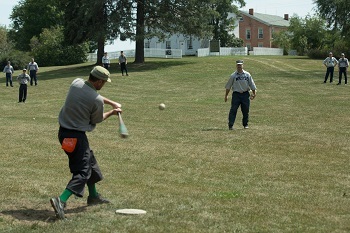A man prepares to hit a baseball just pitched to him, while other field players stand in background. All are dressed in vintage base ball clothes.