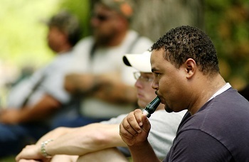 head and shoulders view of a man in a navy blue T-shirt blowing a dark green duck whistle, with other people sitting in background