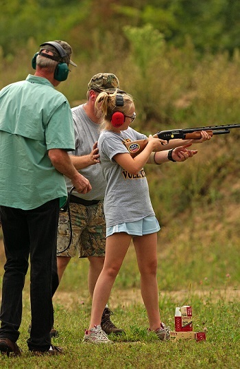 two older men guide a young girl, all wearing ear protection, as she sights in a rifle outdoors