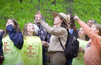 a woman in a tan hat and brown long-sleeved uniform, surrounded by school-age children outdoors, points out something above them