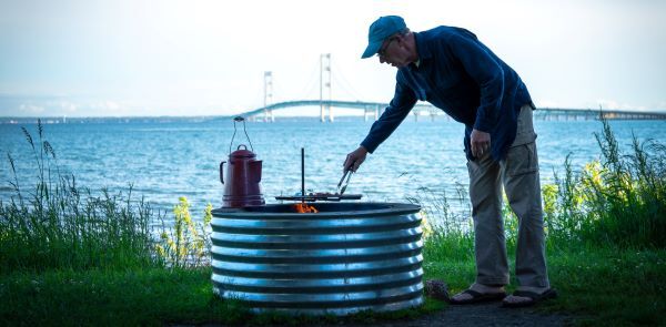 Man cooks hot dogs over a campfire with the Straits of Mackinac and the Mackinac Bridge behind him while at Straits State Park in St. Ignace.