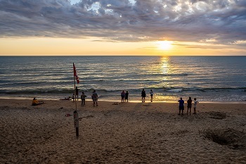 several people outlined in shadow stand on a sandy beach during a golden sunset, as waves roll in. A red beach warning flag is posted