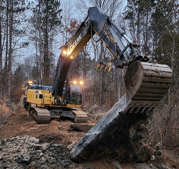 a yellow and black Deer crane with a digging bucket scoops up part of an earthen dam along the rocky shoreline of a shallow body of water