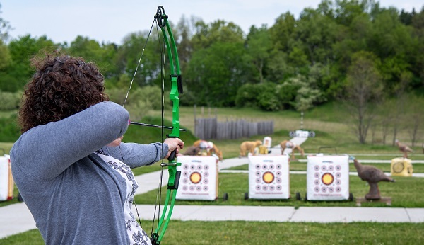 a woman with dark, curly hair pulls back on a neon green bow, aiming her arrow at a row of red and white target bales on the ground