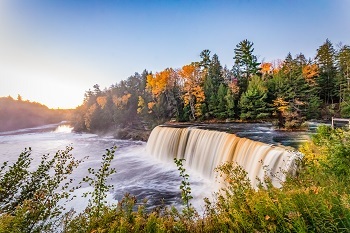 wide waterfall of dark green and rust colored water spills into a curving river; the area is full of mature trees of gold, green and russet