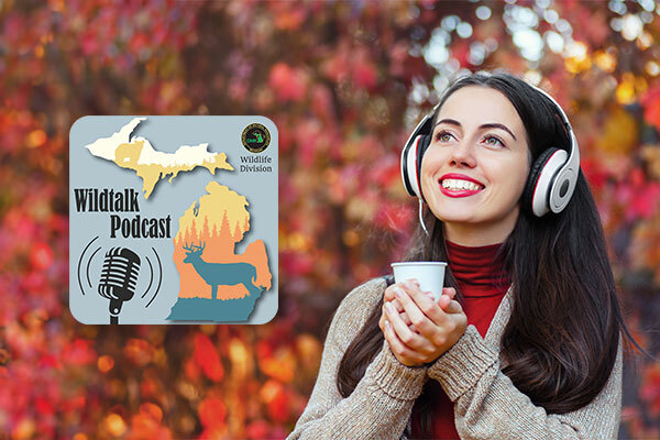 A smiling woman stands in the fall scenery with a sweater, headphones and a steaming hot beverage. 