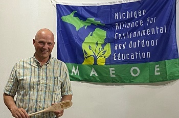 a bald, smiling man in a pale, striped shirt holds a miniature etched wooden oar, in front of a blue and green banner on the wall