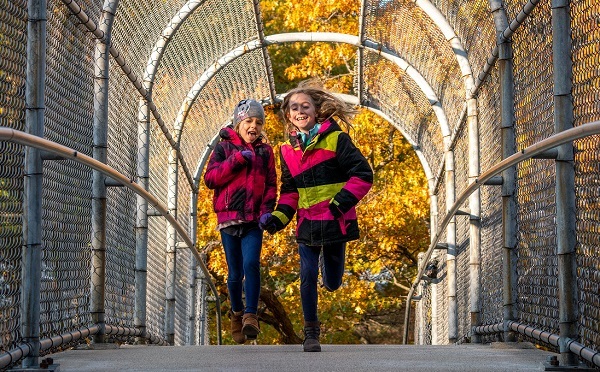 two young girls in brightly colored jackets and jeans run across a fence-covered bridge crosswalk, with bright gold autumn trees behind them