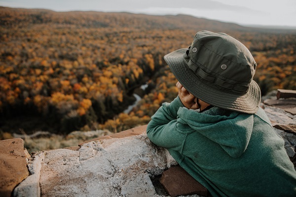 a young child in a teal sweatshirt and green brimmed hat leans his face on his hands, staring out at the vast orange, gold forest below