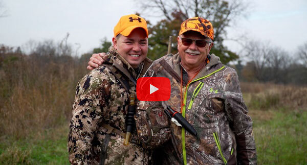 a young man and older man, both smiling and in green camo hunting gear and orange caps, in a grassy field. A play button is overlaid.