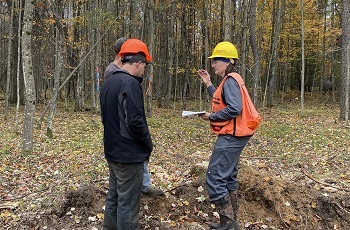 a woman in orange safety vest and yellow hardhat talks with two men in orange hardhats, standing in a muddy forest clearing
