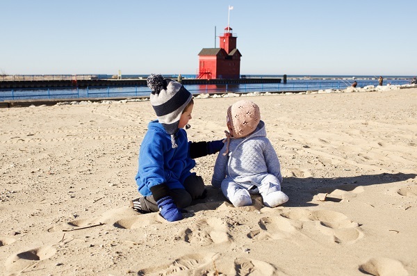Little boy in blue jacket and knit cap with his arm around a toddler girl in quilted snowsuit and peach cap, on sand in front of red lighthouse