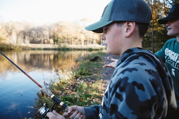 two young boys in baseball caps, and one in camo and one in an MSU Spartan shirt, fish from the shore of a calm, deep blue river