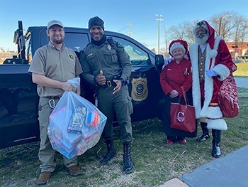 a male park ranger with a bag of toys, a male conservation officer and two people dressed as Santa and Mrs. Claus in front of DNR patrol truck