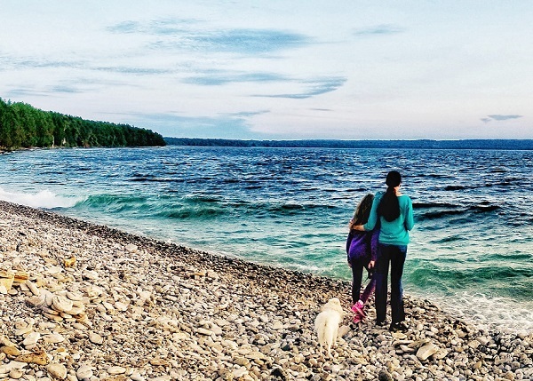 a dark-haired woman and a dark-haired girl stand side by side on a rocky shore, looking out over a large teal and deep blue lake