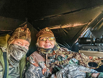 Two women wearing camouflage clothing sit in a hunting blind.