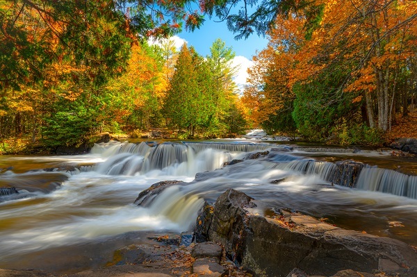 Dark green, foamy water spills over several levels of shallow, wide, rocky falls, with bright green, gold and orange-hued trees in background