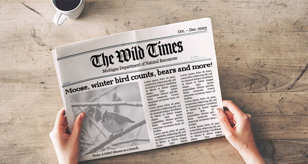 A human hand holds a black and white newspaper with the headline "Moose, winter bird counts, bears and more!"