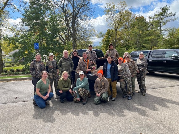 A group of sixteen women dressed in hunting clothes stand together.