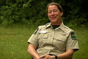 a smiling woman, her brown hair pulled back, in a khaki short-sleeved work shirt with a DNR patch, sits outside in a forested area