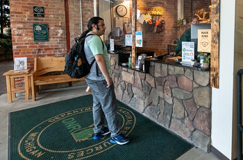 a man in T-shirt, jeans and backpack stands on a green DNR rug, next to a stone welcome counter in a well-outfitted office