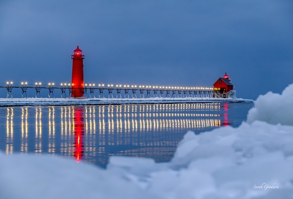 The lit-up pier at Grand Haven State Park, with the two bright red lighthouses against the blue sky; white snow piled in foreground