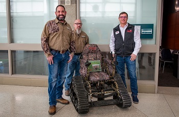 Three smiling men in jeans and long-sleeved shirts flank a camo-colored, wheeled track chair outside a building's concrete, glassed wall