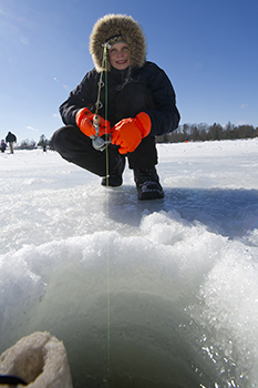 person holding fishing pole on frozen lake with ice fishing hole in foreground