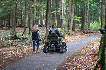 a couple on a paved trail in woods, one person in a track chair