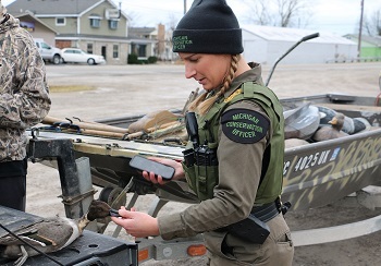 a woman in a DNR conservation officer uniform and hat examines a duck in the back of a pickup while checking a hunter's license info.