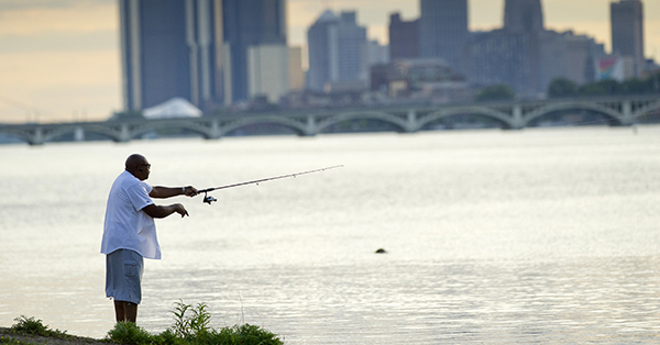 man fishing from bank of Detroit River with city skyline in background