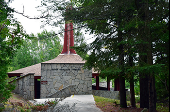 A circular stone pavilion with a shingle roof that comes to a point with four tall red spires is surrounded by pine and cedar trees.  