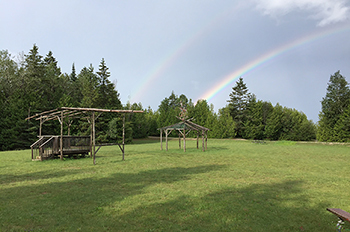 Two structures made from sapling logs sit in the middle of a grassy clearing. A double rainbow extends over pine and cedar trees.