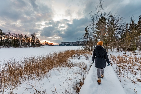 woman in winter gear, orange knit hat and boots walks down a snow-covered, wooden bridge over frozen, snowy waterway. Sun peeks through clouds