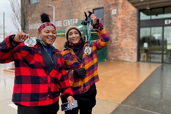 two smiling women, dressed in red and orange flannel, leggings and hats, show off their medals and race bibs in front of a brick building