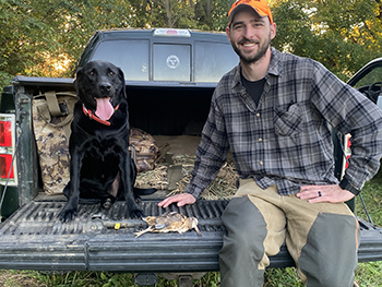 Smiling hunter sits on truck tail bed with black lab, a harvested woodcock in between them