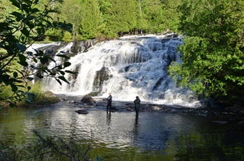 two people in waders and caps stand in shallow water, fishing poles cast, in front of a tall, banked waterfall in a snowy, forested area