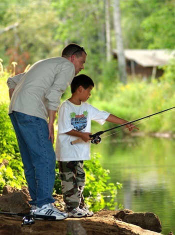 A man helps a young boy fishing from a shaded, rocky area of a riverbank, with sunlight streaming in from above