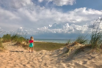 little girl looks stands among sand dunes, looking out at a calm lake, against a bright blue sky dotted with clouds