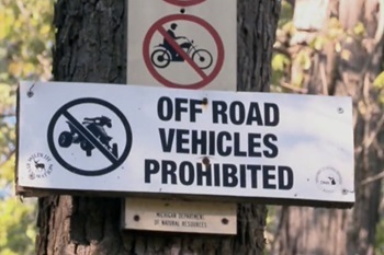 White, black and red signpost saying off-road vehicles prohibited, with circle clash graphics showing same, nailed to a tree in forest