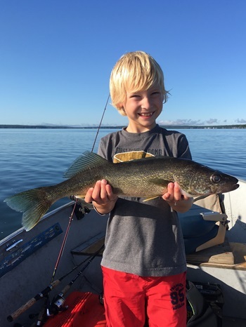 A young, blond child grins while standing in a boat and holding a large walleye. Bright sunshine and blue sky flood the background