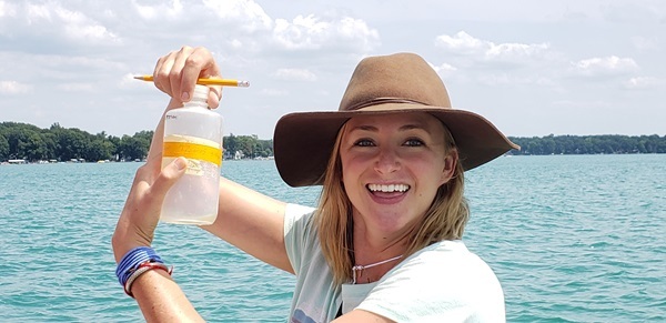 Smiling young woman in a floppy hat and white T-shirt holds a yellow-capped container of water shoulder-high, in front of large lake