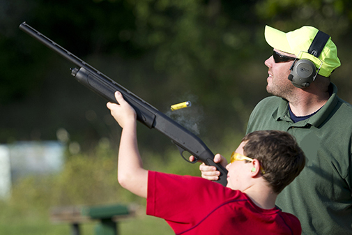 youth shoots shotgun at target with adult looking on