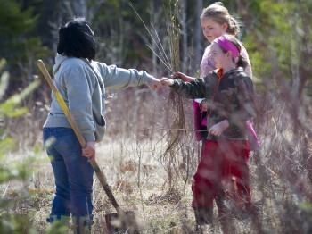 Two children holding bare-root saplings fist-bump an adult volunteer holding a shovel.