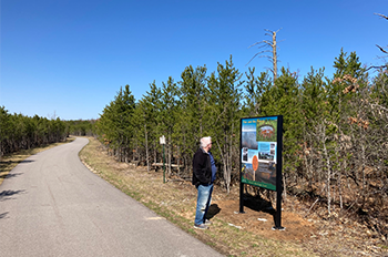 A man reading a Heritage Trail sign along a trail in a wooded area.