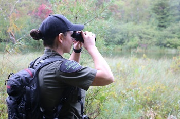 A conservation officer, dark hair twisted in a bun under their baseball cap, gazes through a set of binoculars.